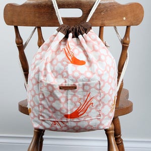 May include: A drawstring backpack featuring a light pink and white clover pattern with orange octopus illustrations. The bag has a brown fabric top closure, white rope straps, and a front pocket with a small rectangular tag labelled HAND MADE.