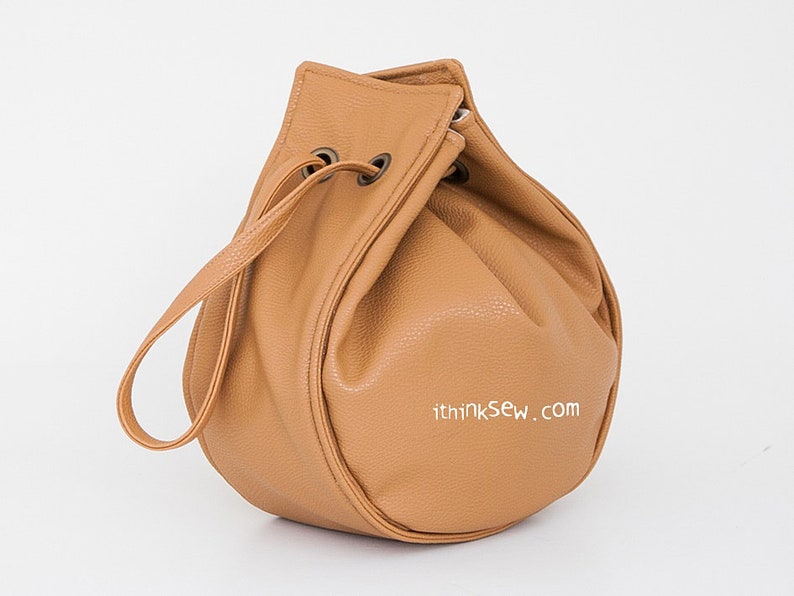 Peut inclure: Un sac seau en cuir brun clair avec une seule sangle. Le sac a une forme ronde et est fait d'un cuir souple et textur&eacute;. Le sac a une adresse de site Web imprim&eacute;e sur le fond : "ithinksew.com".