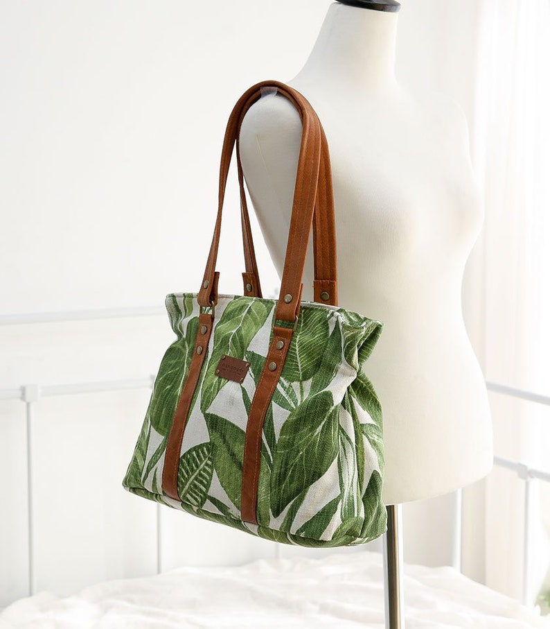 May include: A green and white canvas tote bag with brown leather straps and accents. The bag features a tropical leaf print.