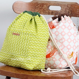 Op de afbeelding: Twee trekkoordtassen, een groene met een geometrisch patroon en een roze met een wit stippenpatroon. Beide tassen hebben een klein vakje aan de voorkant en een wit trekkoord.