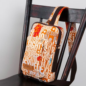May include: A sling backpack featuring an orange and white fabric with a repeating travel-themed typography print. The bag includes a front zippered pocket, a brown strap, and a small rectangular leather-like patch labeled handmade on the front.