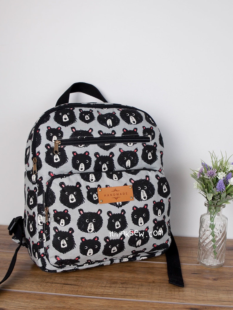 May include: A grey backpack with a black bear print and the text "Handmade" on a brown leather tag. The backpack has black straps and a black zipper.