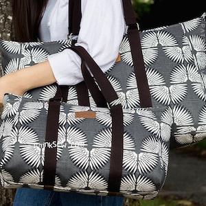 Op de afbeelding: Twee grijze en witte tote bags met bruine banden. De tassen hebben een patroon van witte gestileerde vleugels op een grijze achtergrond. De grotere tas heeft een band die over de schouder gaat. De kleinere tas heeft twee kortere banden. De tassen zijn gemaakt van canvas.