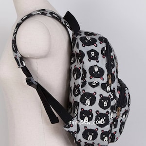 May include: A grey canvas backpack with a black bear print. The backpack has two adjustable straps and a zip closure. The backpack is on a mannequin.