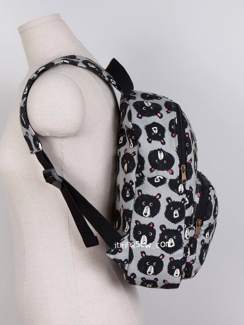 May include: A grey canvas backpack with a black bear print. The backpack has two adjustable straps and a zip closure. The backpack is on a mannequin.