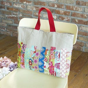 Puede incluir: Una bolsa de lona grande de color beige con asas rojas. La bolsa está hecha de un mosaico de tiras de tela de colores con patrones florales y geométricos. La bolsa está sentada sobre una superficie de madera marrón claro.