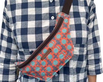 May include: A handmade fanny pack with a floral pattern in orange and teal on a gray background. The bag has a brown zipper and strap, and a small "handmade" tag. The person is wearing a blue and white checkered shirt.