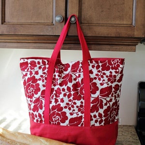 May include: A red and white floral tote bag with a solid red bottom. The bag has two long straps and is hanging from a cabinet door. Two loaves of bread are on a countertop in front of the bag.