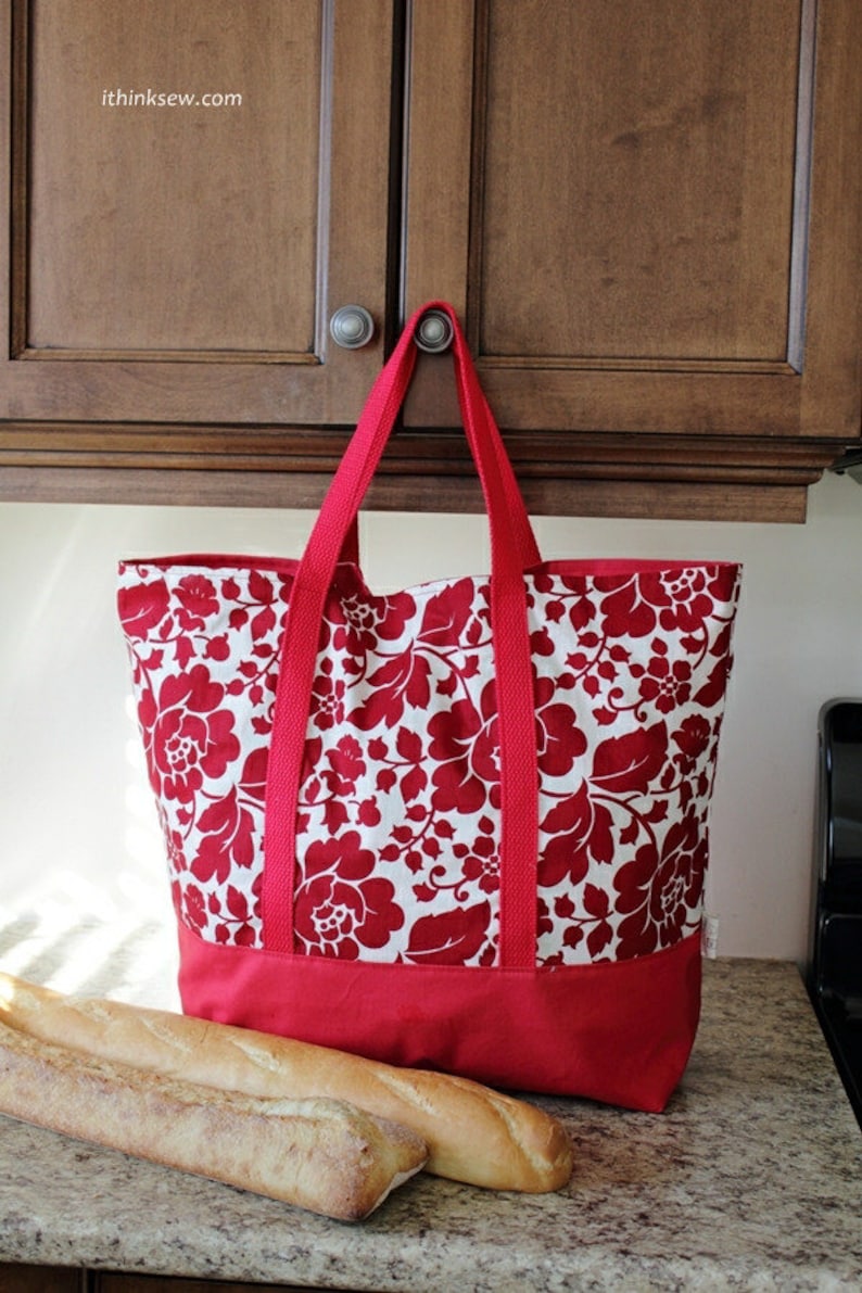 May include: A red and white floral tote bag with a solid red bottom. The bag has two long straps and is hanging from a cabinet door. Two loaves of bread are on a countertop in front of the bag.