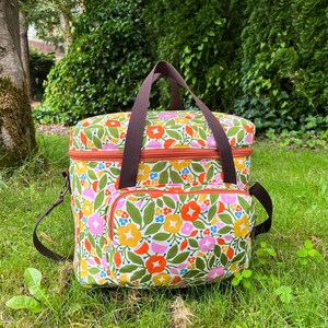 May include: A floral cooler bag with a brown handle and shoulder strap. The bag features a colourful design of flowers in orange, pink, yellow, and blue on a white background. The bag has an orange zip and a front pocket.