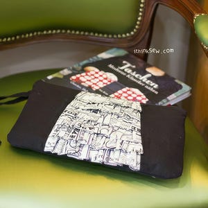 May include: A black fabric clutch with a white and black patterned flap. The clutch is sitting on a green chair with a book titled "Taschen" on top of it.