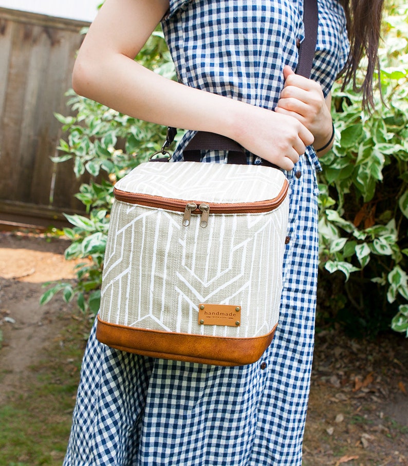 May include: A beige and white geometric patterned insulated lunch bag with a brown leather trim and a brown adjustable shoulder strap. The bag has a zipper closure and a brown leather tag that says "handmade".