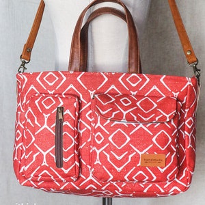 K&ouml;nnte beinhalten: Rote Canvas-Tragetasche mit geometrischem Muster und braunen Ledergriffen. Die Tasche hat zwei Rei&szlig;verschlussf&auml;cher auf der Vorderseite und einen abnehmbaren Schultergurt. Die Tasche hat ein "handgefertigt mit Liebe"-Etikett auf der Vorderseite.