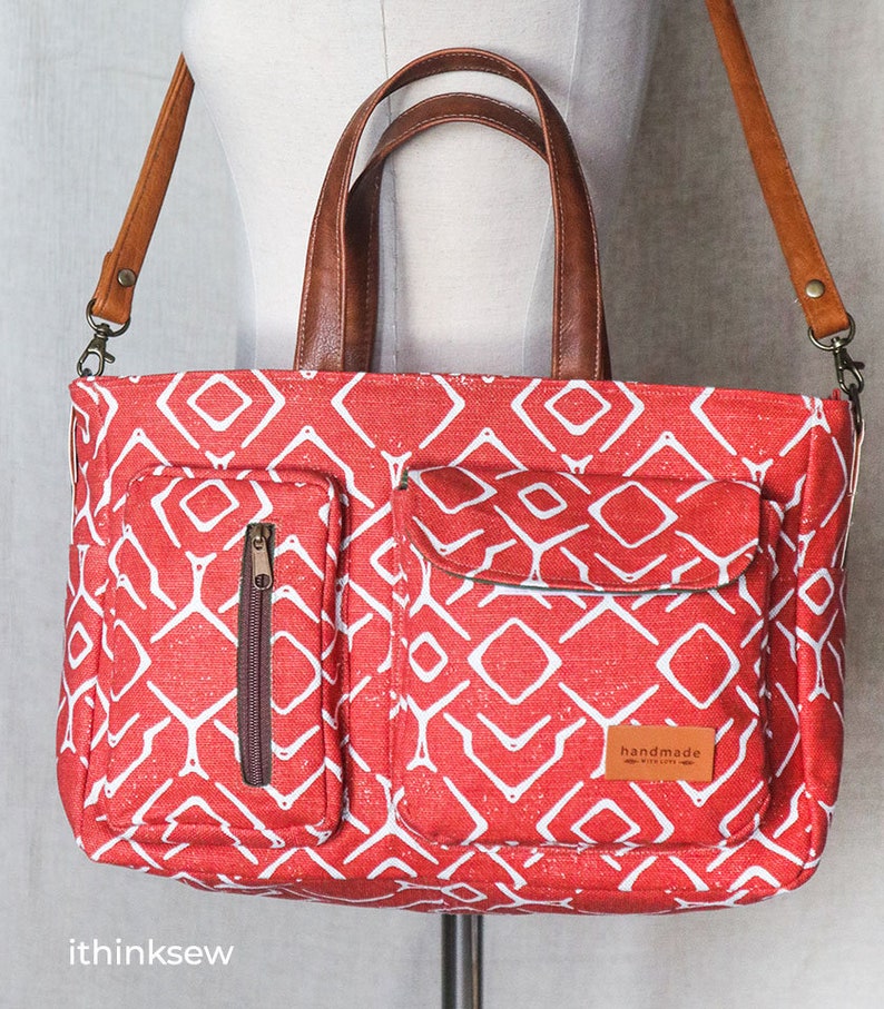 K&ouml;nnte beinhalten: Rote Canvas-Tragetasche mit geometrischem Muster und braunen Ledergriffen. Die Tasche hat zwei Rei&szlig;verschlussf&auml;cher auf der Vorderseite und einen abnehmbaren Schultergurt. Die Tasche hat ein "handgefertigt mit Liebe"-Etikett auf der Vorderseite.