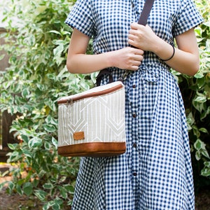 May include: A white and brown lunch bag with a geometric pattern and a brown leather trim. The bag has a zipper closure and a black shoulder strap.