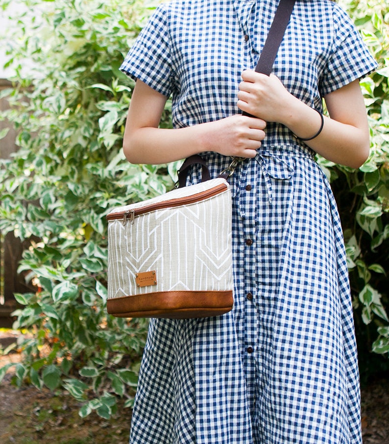 May include: A white and brown lunch bag with a geometric pattern and a brown leather trim. The bag has a zipper closure and a black shoulder strap.