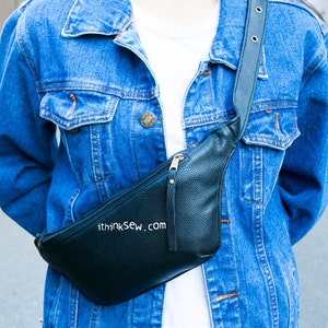 May include: A black leather fanny pack with a zipper closure and a gold zipper pull. The fanny pack is worn over a denim jacket.