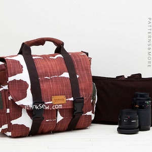 May include: A brown and white patterned camera bag with a brown leather handle and a green zippered pocket. The bag has a brown adjustable strap and a brown leather label that says "Handmade ithinksew.com". The bag is sitting on a white surface with two camera lenses in front of it.