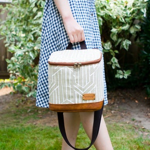 May include: A beige and brown insulated lunch bag with a geometric pattern and a brown leather strap. The bag has a brown leather label that says "handmade".