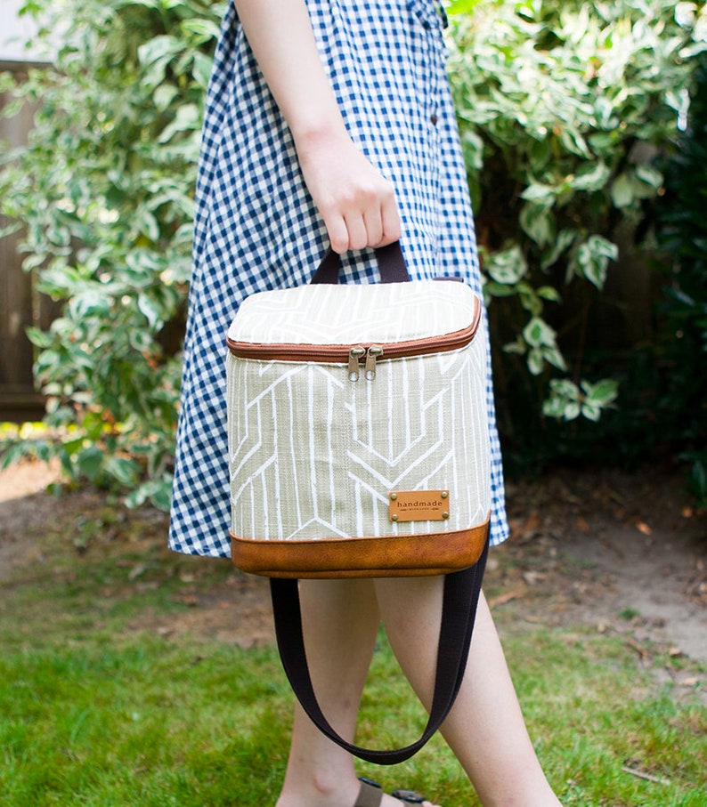 May include: A beige and brown insulated lunch bag with a geometric pattern and a brown leather strap. The bag has a brown leather label that says "handmade".