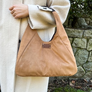 May include: A tan suede shoulder bag with a brown leather handle and a brown leather patch that says "handmade".