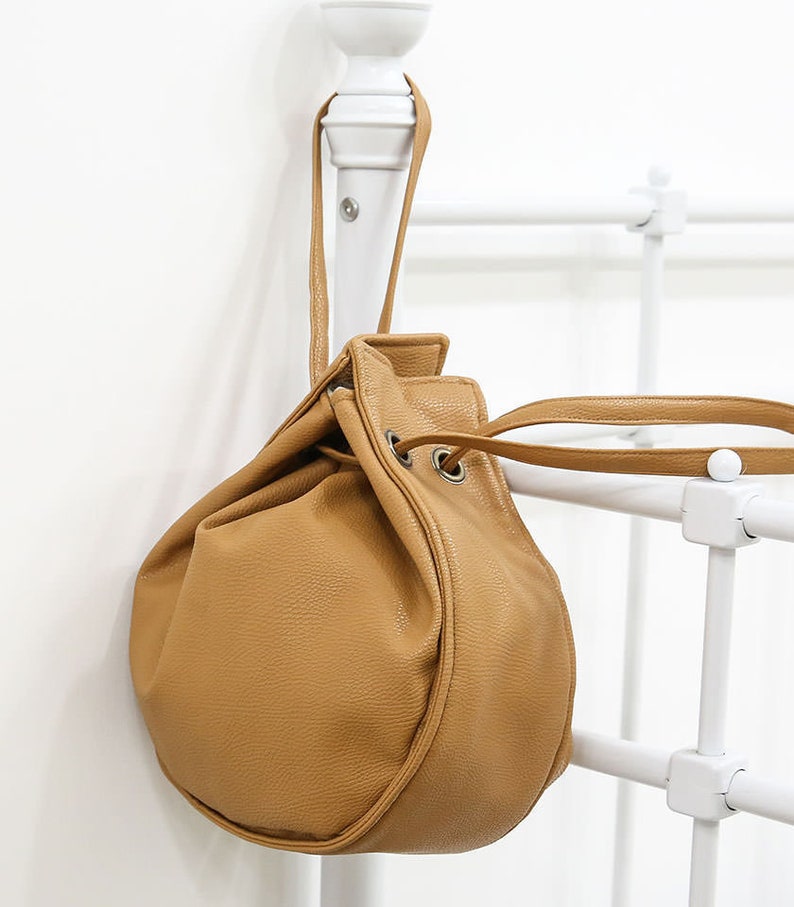 Peut inclure: Un sac seau en cuir marron avec une fermeture &agrave; cordon et une longue bandouli&egrave;re. Le sac est suspendu &agrave; un support en m&eacute;tal blanc.
