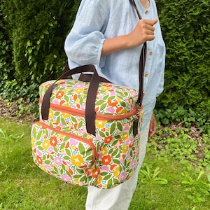 May include: A shoulder bag with a floral pattern, brown straps, and orange trim. The bag displays a colourful design of flowers and leaves in yellow, pink, and green. It has a front pocket and a top zip closure.