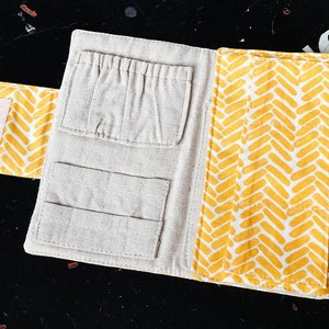 Könnte beinhalten: Ein beigefarbener Stofforganizer mit mehreren Taschen und einem gelben Chevron-Muster auf der Außenseite. Der Organizer ist geöffnet und die Taschen sind sichtbar. Der Text "ROBIN + BIRCH" ist auf der rechten Seite des Organizers aufgedruckt.