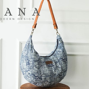 Peut inclure: Un sac cabas bleu et blanc à motifs avec une bandoulière en cuir marron. Le sac a une forme incurvée et un logo DANA MODERN DESIGN.
