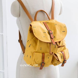 Pode incluir: Uma mochila de lona amarela mostarda com alças e fivelas de couro castanho. A mochila tem dois bolsos frontais com fivelas e um fecho de cordão no topo. A mochila tem um pequeno padrão preto e branco.