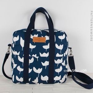 May include: A blue canvas tote bag with white tulip print and navy blue straps. The bag has a brown leather label with the text "Handmade" on it. The bag has a long adjustable strap for carrying it on the shoulder.