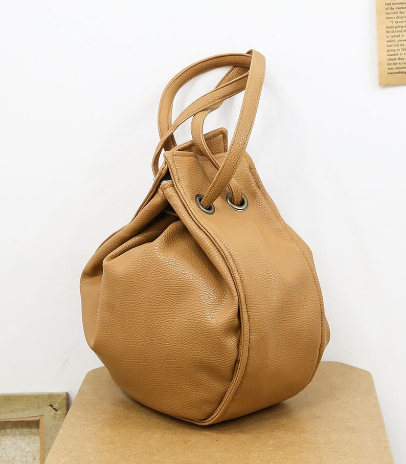 Peut inclure: Un sac seau en cuir de couleur tan avec une fermeture &agrave; cordon et deux poign&eacute;es sup&eacute;rieures. Le sac a une forme ronde et l&eacute;g&egrave;rement affaiss&eacute;e.