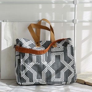 May include: A large gray and white geometric patterned tote bag with brown leather handles and a shoulder strap. The bag has a zippered pocket on the front.