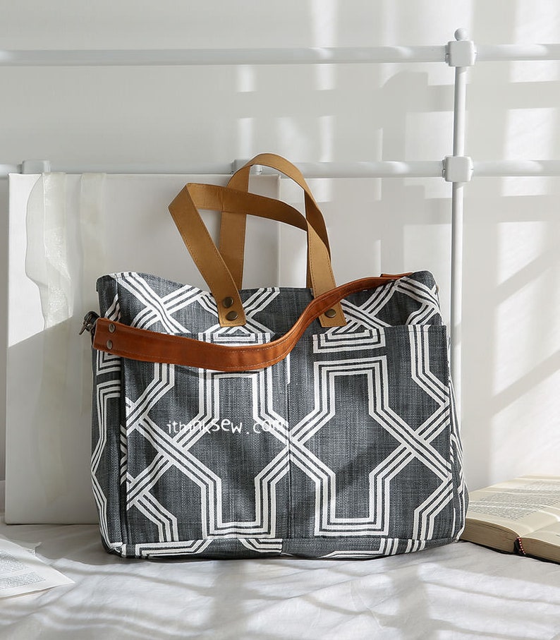 May include: A large gray and white geometric patterned tote bag with brown leather handles and a shoulder strap. The bag has a zippered pocket on the front.