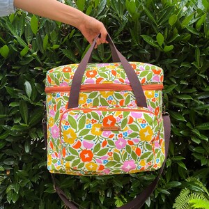 May include: A colourful floral cooler bag with brown straps and handles. The bag displays a pattern of orange, pink, yellow, and blue flowers with green leaves on a white base. It has a front pocket and a brown zip.