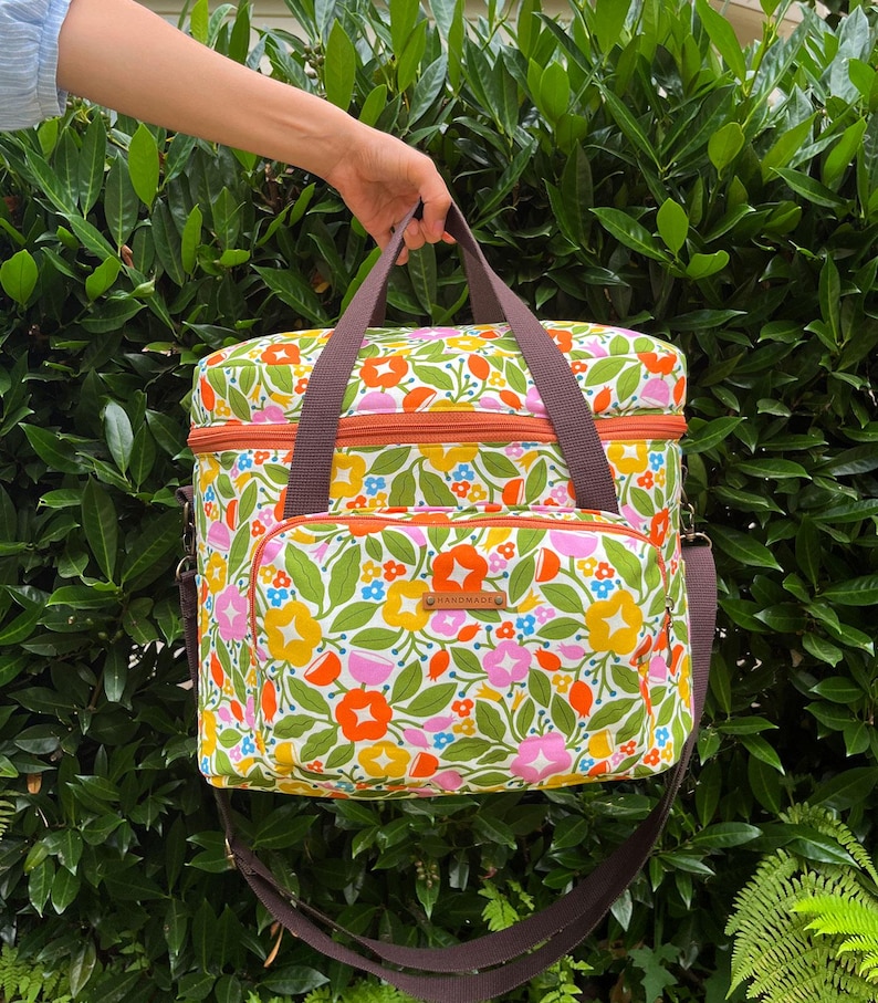 May include: A colourful floral cooler bag with brown straps and handles. The bag displays a pattern of orange, pink, yellow, and blue flowers with green leaves on a white base. It has a front pocket and a brown zip.