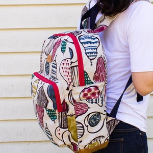 May include: A white canvas backpack with a colorful fruit print. The backpack has red zipper accents and a brown leather bottom. The backpack is being worn by a person.