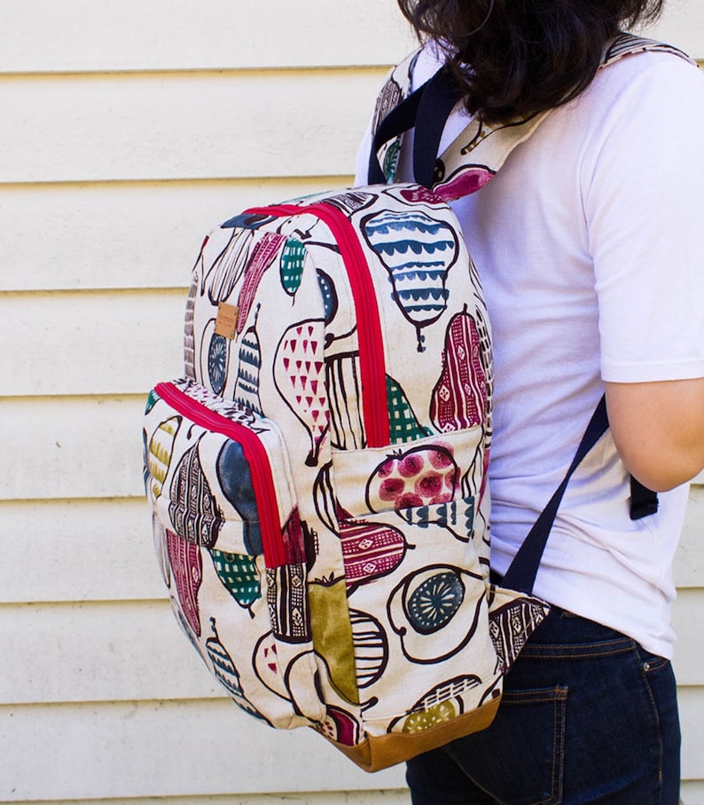 May include: A white canvas backpack with a colorful fruit print. The backpack has red zipper accents and a brown leather bottom. The backpack is being worn by a person.
