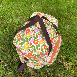 May include: A floral-patterned cooler bag with brown straps and zip trim, set on green grass. The bag features a vibrant design of orange, yellow, pink, and blue flowers with green leaves on a white background.