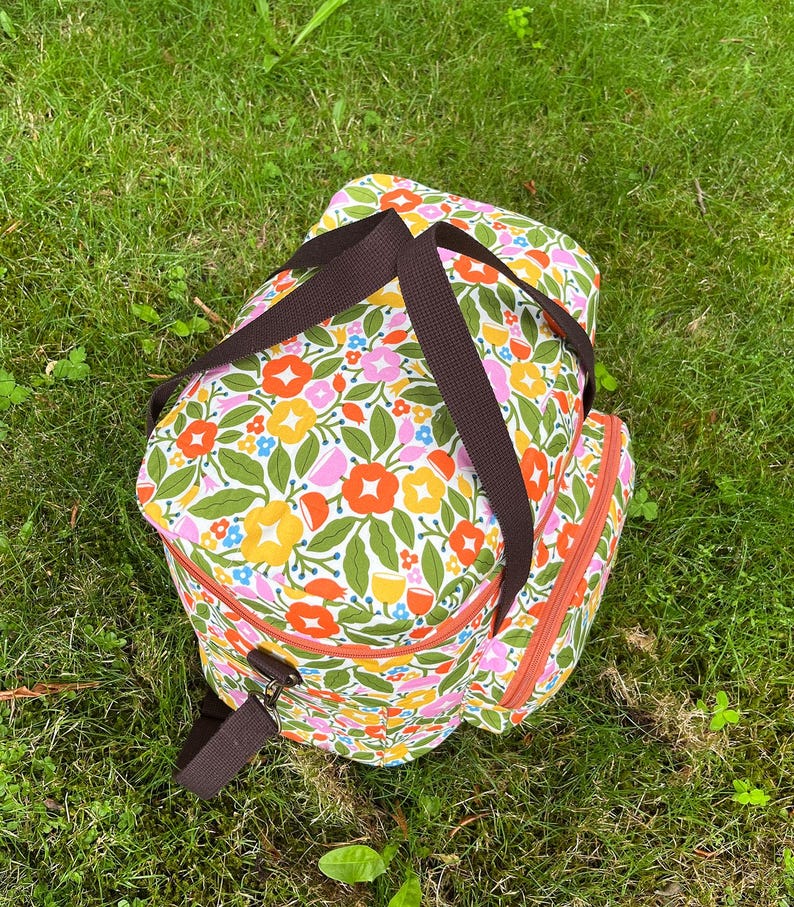 May include: A floral-patterned cooler bag with brown straps and zip trim, set on green grass. The bag features a vibrant design of orange, yellow, pink, and blue flowers with green leaves on a white background.