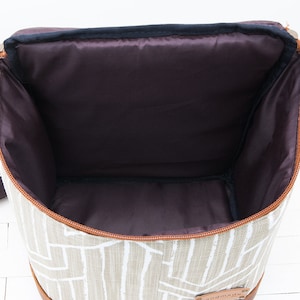 May include: A beige canvas lunch bag with a brown leather trim and a zipper closure. The bag has a white geometric pattern and a brown leather tag that says "Handmade".