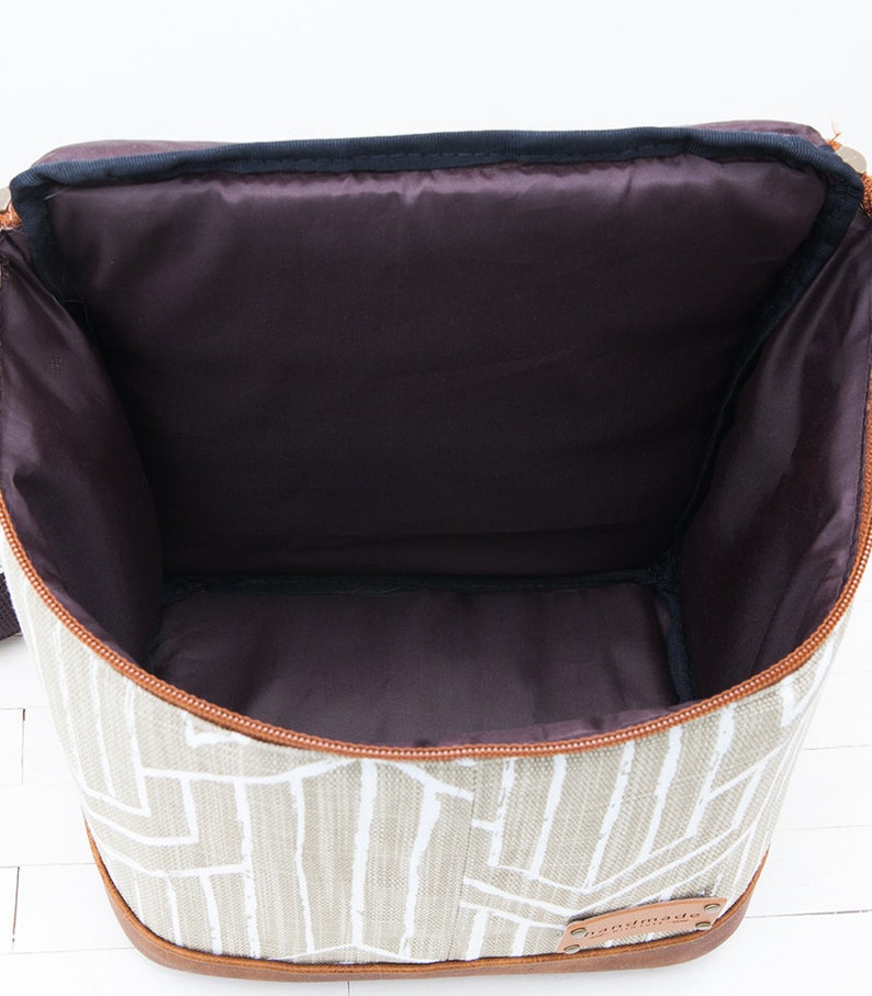May include: A beige canvas lunch bag with a brown leather trim and a zipper closure. The bag has a white geometric pattern and a brown leather tag that says "Handmade".
