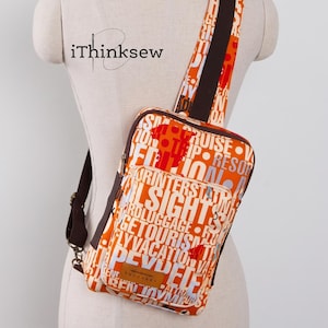 May include: A crossbody sling bag featuring a vibrant orange fabric with a white and dark brown typographic print of travel-related words. The bag includes a dark brown adjustable strap, a front zippered pocket, and a rectangular brown leather label.