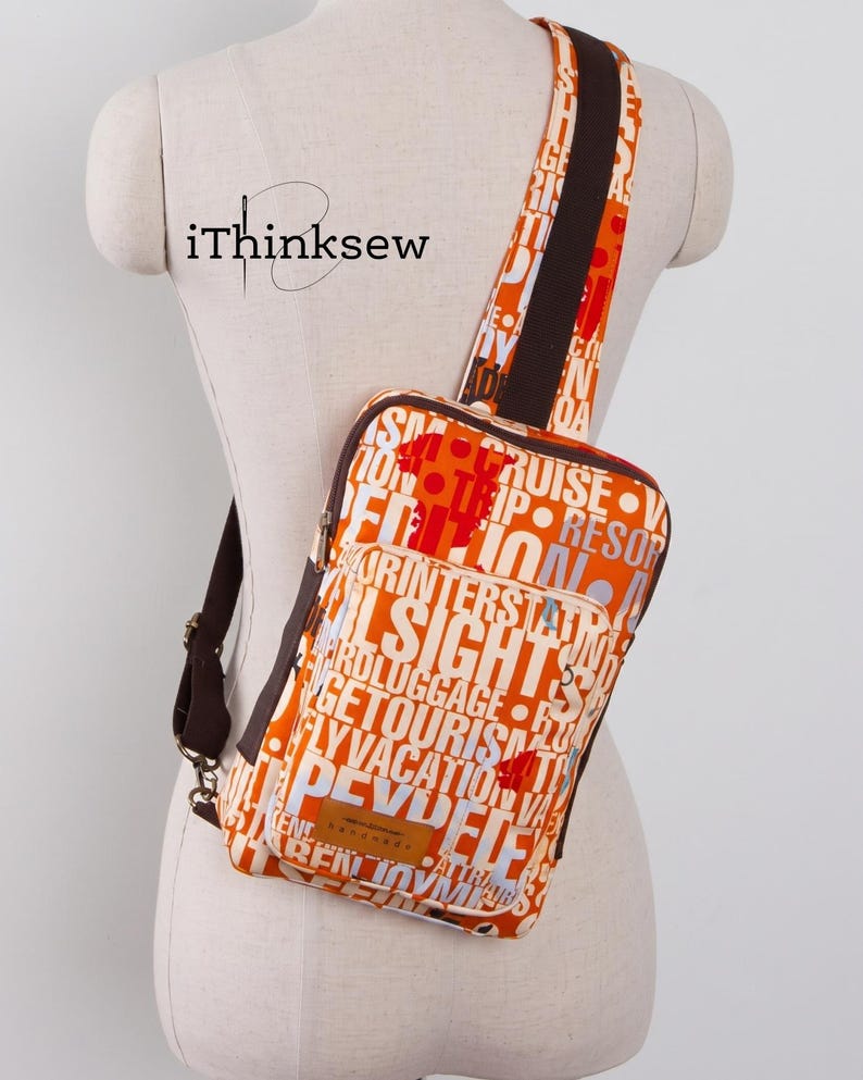 May include: A crossbody sling bag featuring a vibrant orange fabric with a white and dark brown typographic print of travel-related words. The bag includes a dark brown adjustable strap, a front zippered pocket, and a rectangular brown leather label.