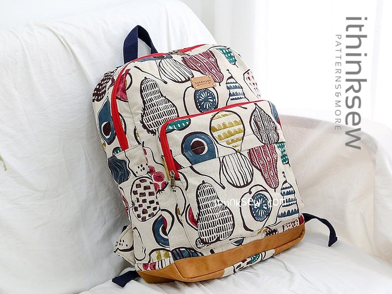 May include: A white canvas backpack with a red zipper and a colorful abstract pattern of pears and other shapes. The backpack has a brown leather bottom and a blue adjustable strap.