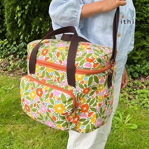 May include: A floral print tote bag with brown handles and a shoulder strap. The bag features a colorful pattern of flowers in orange, yellow, pink, and green. It has an orange zip and a front pocket.