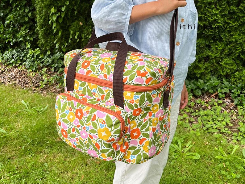 May include: A floral print tote bag with brown handles and a shoulder strap. The bag features a colorful pattern of flowers in orange, yellow, pink, and green. It has an orange zip and a front pocket.