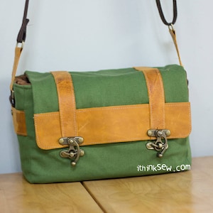 Pu&ograve; includere: Una borsa a tracolla in tela verde con dettagli in pelle marrone e fibbie metalliche. La borsa ha una tracolla e un indirizzo del sito web stampato sul fondo.
