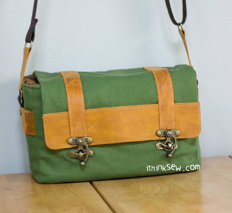 Pu&ograve; includere: Una borsa a tracolla in tela verde con dettagli in pelle marrone e fibbie metalliche. La borsa ha una tracolla e un indirizzo del sito web stampato sul fondo.