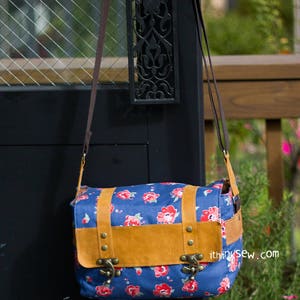 Pu&ograve; includere: Una borsa a tracolla in tela blu con una patta in pelle marrone e fibbie metalliche. La borsa ha una stampa floreale con fiori rossi e dettagli bianchi. La borsa &egrave; appesa a una porta nera con un design in metallo.
